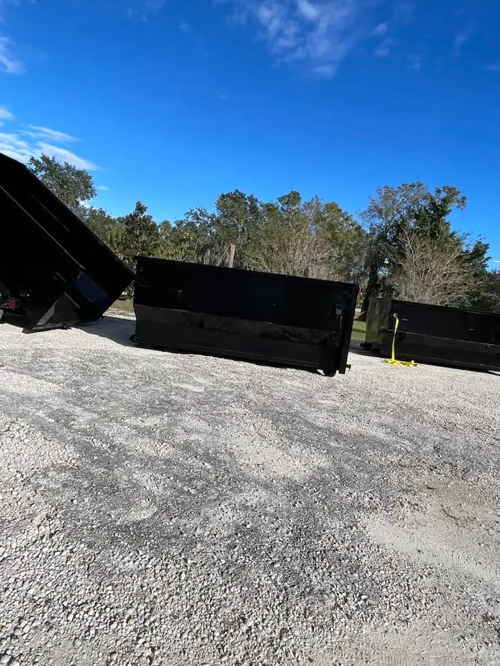 Several large black metal containers are placed on a gravel lot outdoors, with trees and a blue sky in the background.