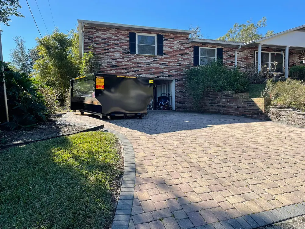 A black dumpster sits on the brick driveway in front of a two-story brick house with a garage and shrubbery on either side.