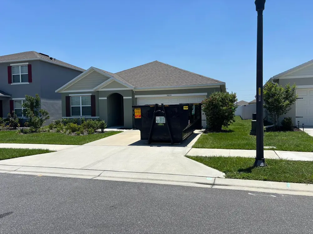 A black dumpster sits in the driveway of a single-story suburban house on a sunny day.