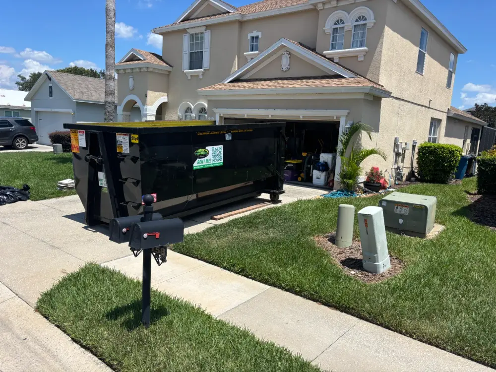 A large black residential dumpster sits in the driveway of a beige suburban house with an open garage and various items scattered on the lawn and driveway.