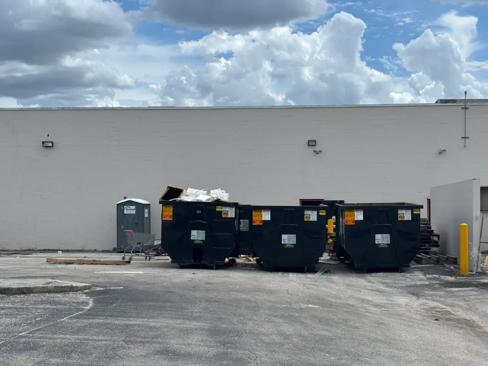 Three large residential dumpsters and a portable toilet are placed near a blank white wall in a parking lot under a partly cloudy sky.