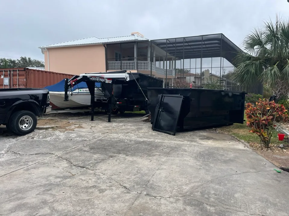 A black dump trailer with an open door is parked on a cracked driveway near a black truck, a boat, and a two-story house with a screened porch.