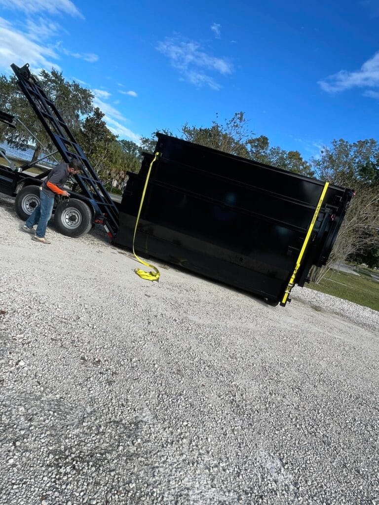 A large black dumpster is tilted on its side near a gravel area, with a man standing nearby and a yellow strap lying on the ground.