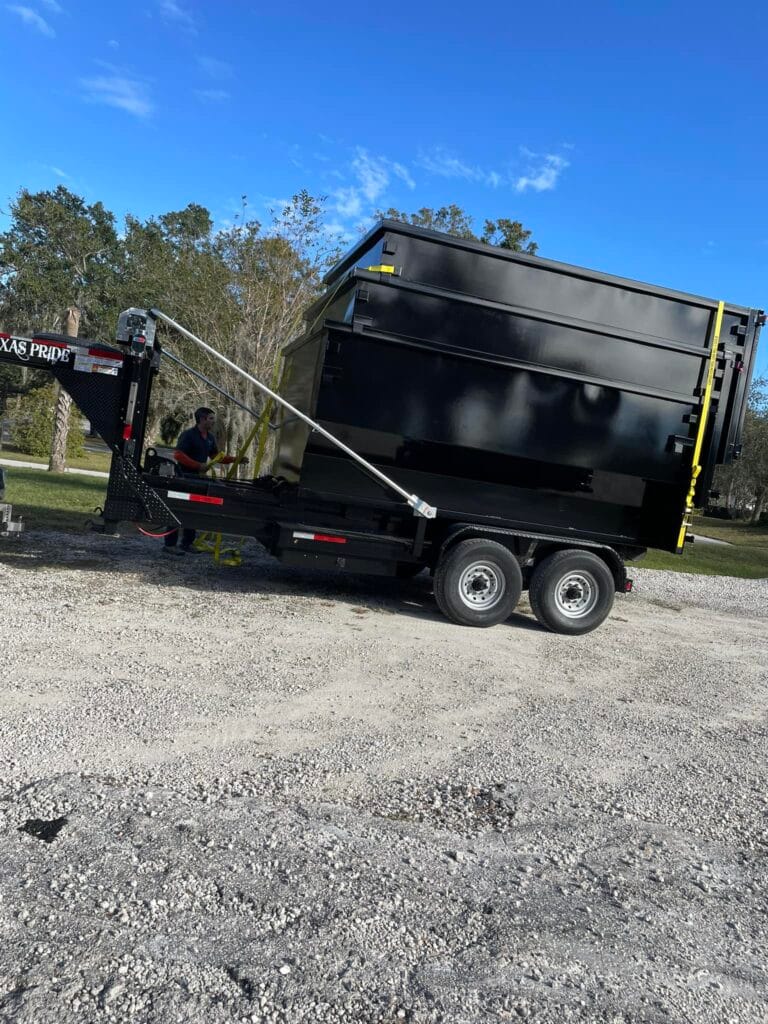 A large black dumpster is loaded on a trailer with dual axles, parked on a gravel area under a clear blue sky. A person is seated near the trailer hitch.