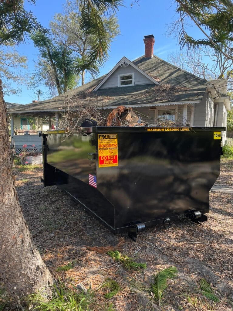 A large black dumpster sits on the front lawn of a house, with trees and yard debris visible around it on a sunny day.