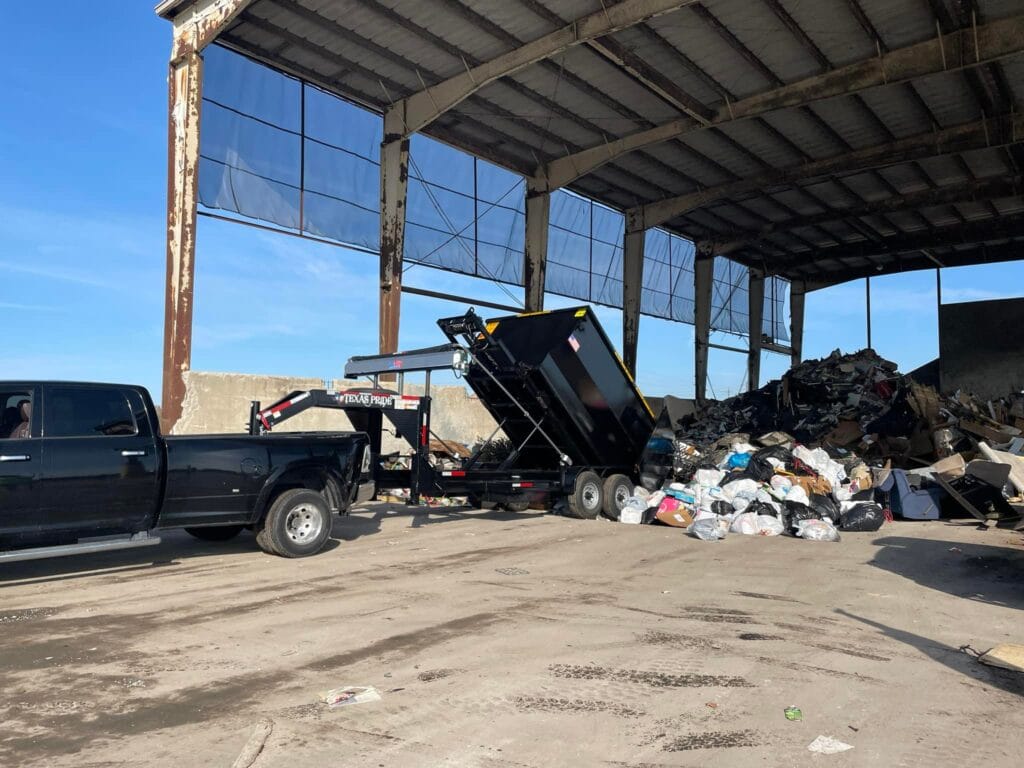 A black truck with a trailer is dumping garbage at a waste disposal facility inside a large, partially open building with piles of trash.
