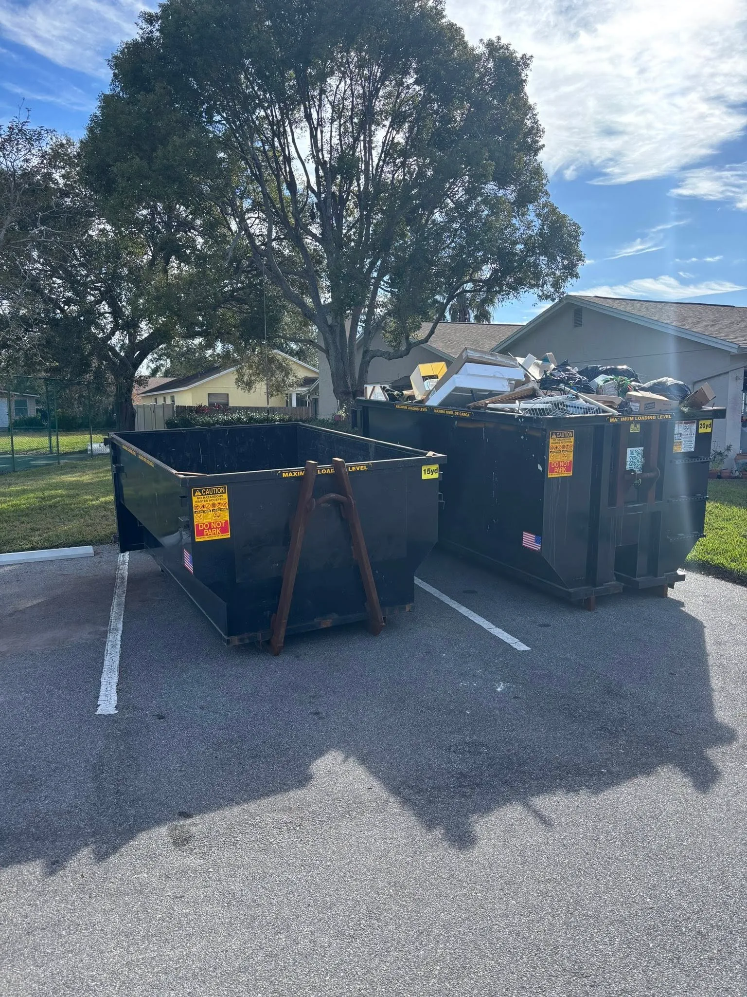 Two large dumpsters in a parking lot, one empty and one filled with debris and discarded items, with houses and trees in the background on a sunny day.