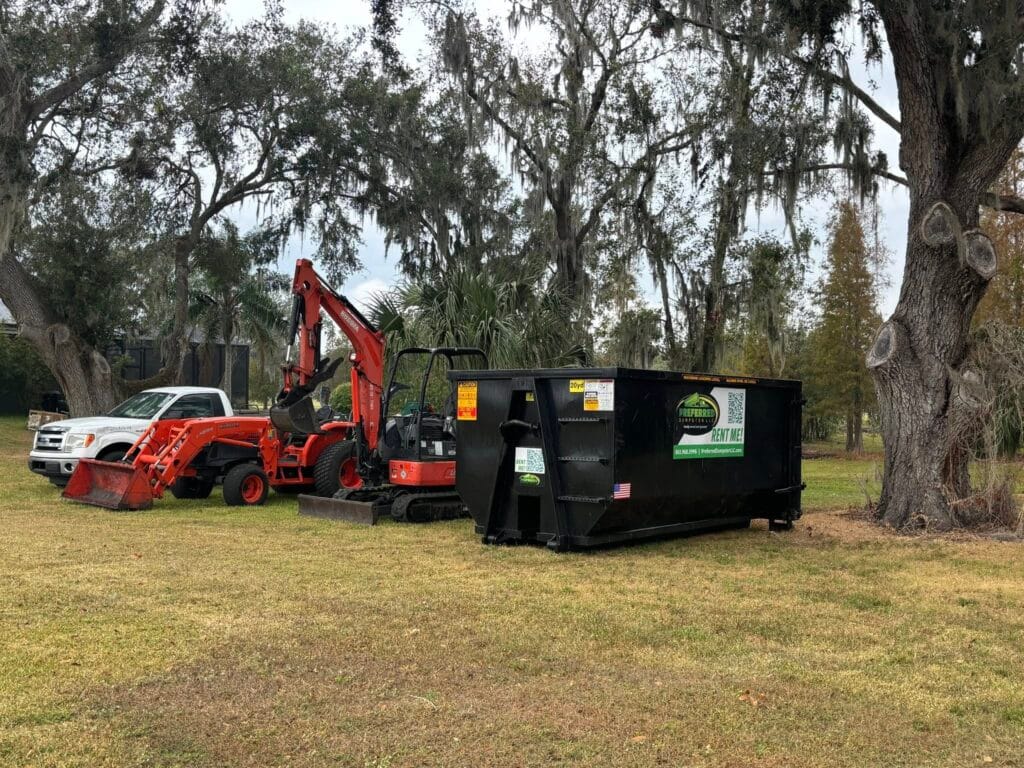A white pickup truck, a red excavator, and a black dumpster are parked on a grassy area near trees with Spanish moss.