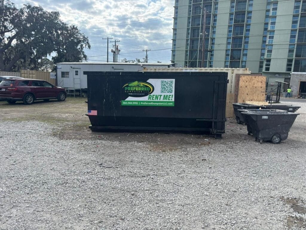 A large black dumpster with a "Preferred" rental sign is placed on a gravel lot near a building and parked vehicles.