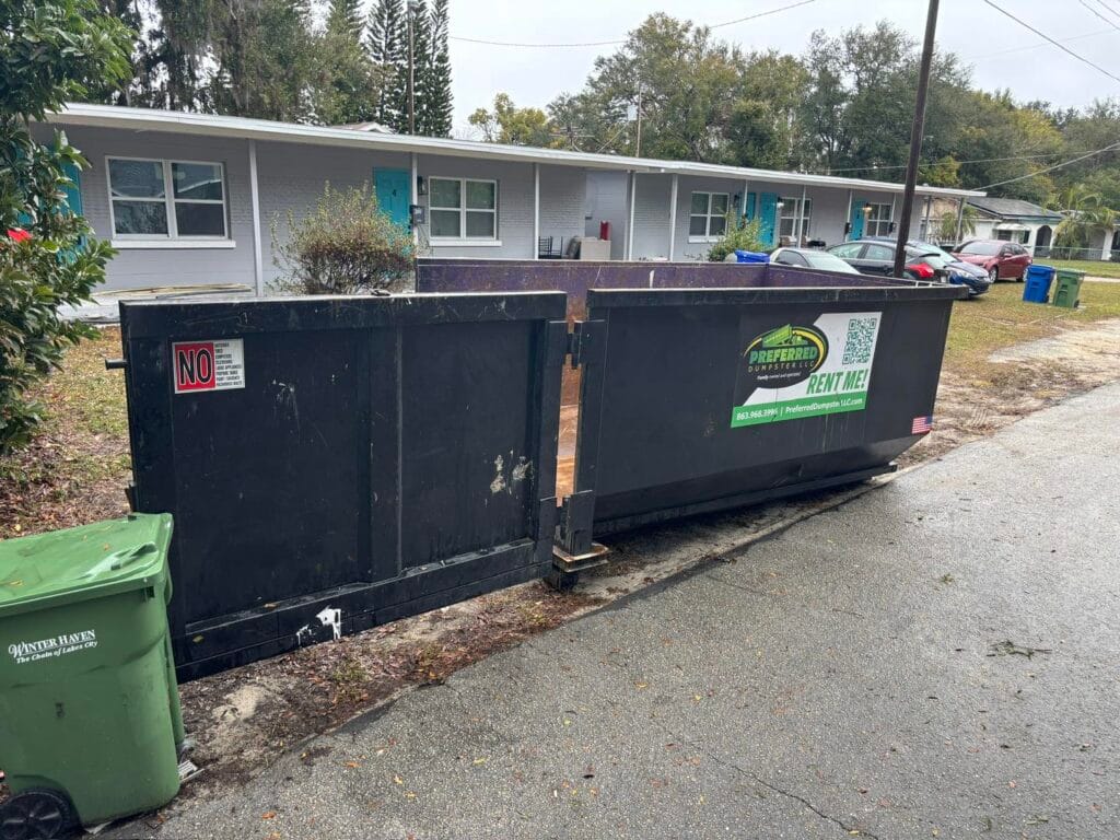 A large black dumpster and a green trash bin are positioned on the street in front of a row of single-story houses on a cloudy day.