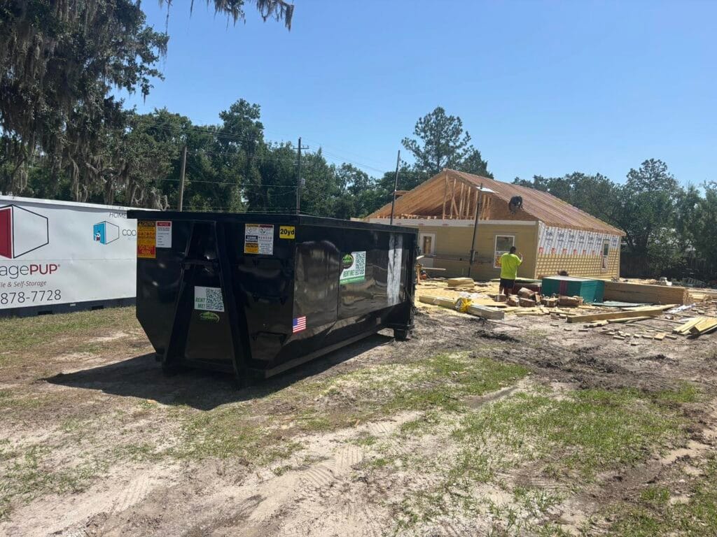 A construction site with a large black dumpster in the foreground and a worker near a partially built house in the background.