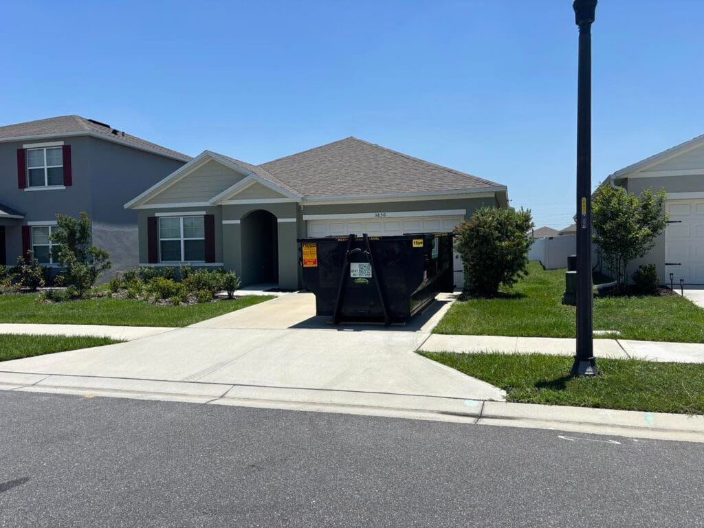 A black dumpster sits in the driveway of a single-story suburban house on a sunny day.