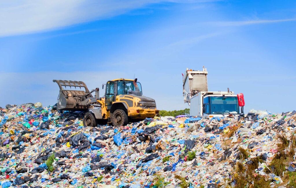 A bulldozer moves through a large landfill covered with plastic and other waste under a blue sky.