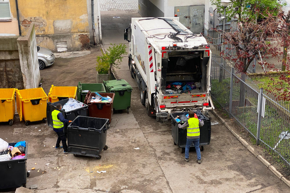 Two sanitation workers in yellow vests empty large garbage bins into a white garbage truck in an urban alley, surrounded by yellow and green bins—similar to those used for yard waste dumpster rental services.