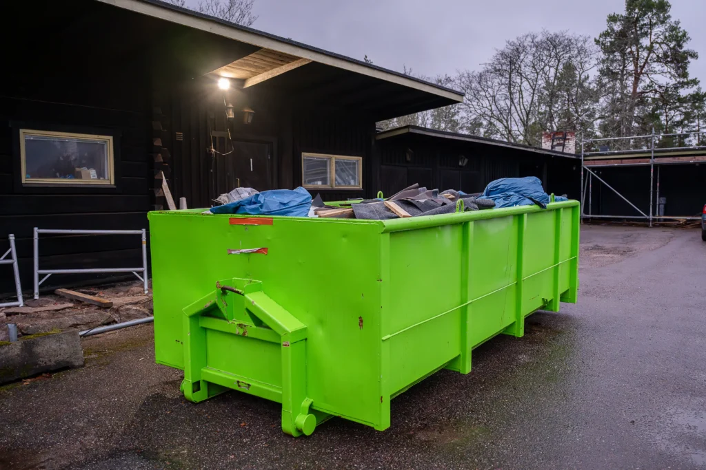 A large, bright green dumpster filled with assorted debris sits outside a dark building on a wet, overcast day, highlighting the importance of knowing what can you throw in a dumpster.