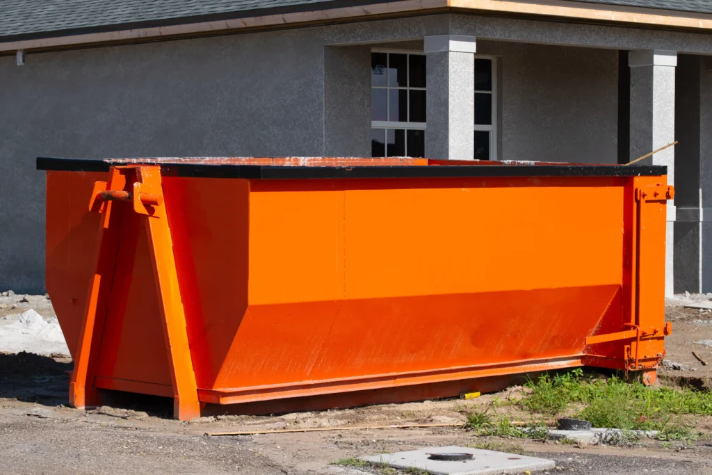 A large orange metal dumpster sits on a paved area in front of a gray building with white trim and a window.