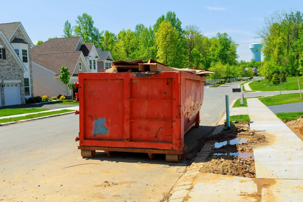 A large red dumpster sits on the street in a suburban neighborhood, surrounded by houses and trees, with debris and muddy water on the ground nearby.