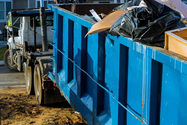 A large blue dumpster filled with various trash items is positioned on a construction site beside a white truck.