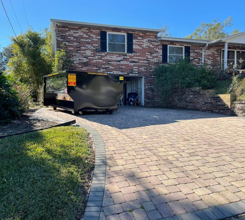 A black dumpster sits on the brick driveway in front of a two-story brick house with a garage and shrubbery on either side. A black dumpster sits on the brick driveway in front of a two-story brick house with a garage and shrubbery on either side.