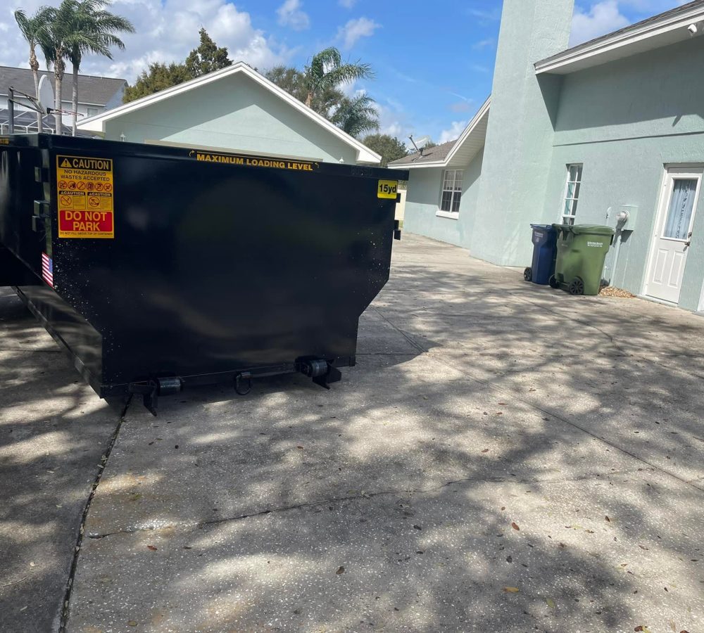 A large black dumpster with caution signs is placed on the driveway of a light blue house with palm trees in the background. A large black dumpster with caution signs is placed on the driveway of a light blue house with palm trees in the background.