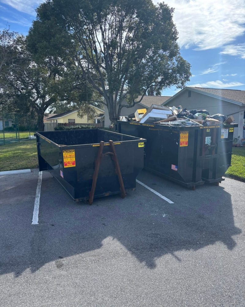 Two large dumpsters in a parking lot, one empty and one filled with debris and discarded items, with houses and trees in the background on a sunny day.